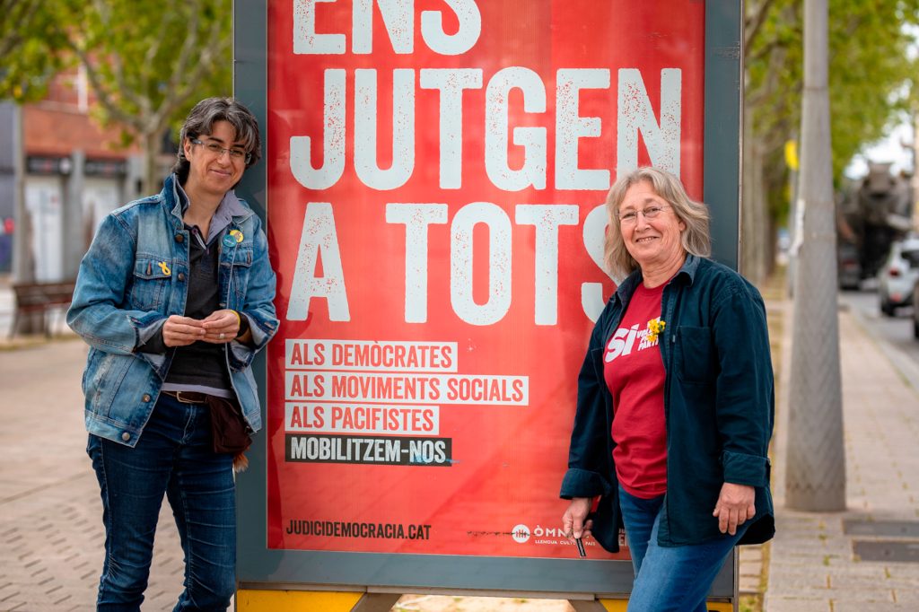 L'Eva Navarrete i la Maria Forns davant d'un cartell d'Òmnium durant la campanya de les Municipals 2019, que diu "Ens jutgen a tots".